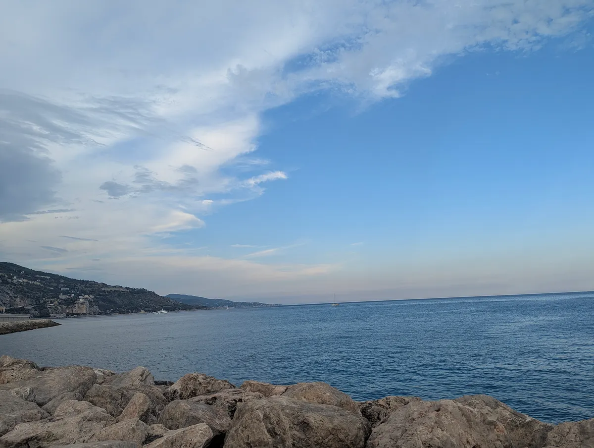The sea under a wide blue sky with wispy clouds, seen from a rocky shore, with a hilly coastline in the distance.