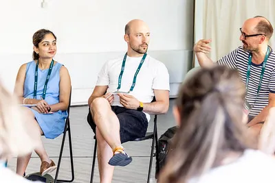 A small group discussion at a conference: three people seated on chairs, one speaking with a gesture, while others listen.